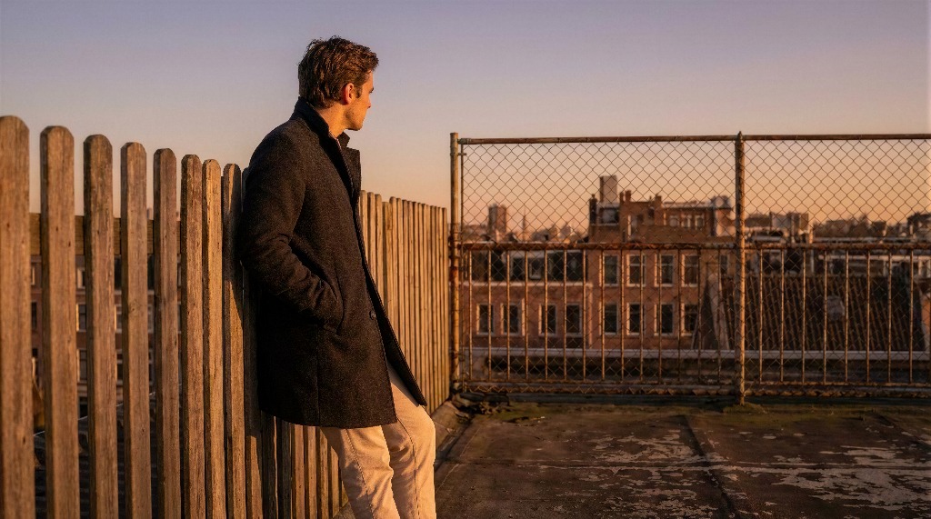 Man in a coat gazes at cityscape from a rooftop at sunset.