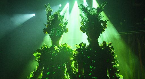 Two performers in elaborate green costumes under stage lights