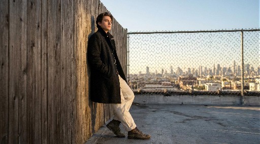 Young man in a coat leans against a wooden fence on a rooftop.