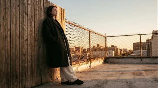 Young woman in a coat leans against a rooftop fence at sunset.
