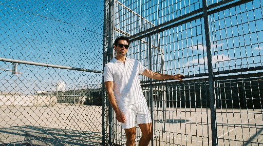 Man in white outfit leaning on a chain-link fence under a clear blue sky.